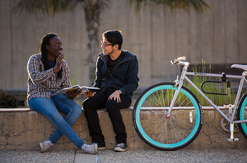 Image of two students discussing the textbook while sitting on a ledge, with bike perched against the edge
