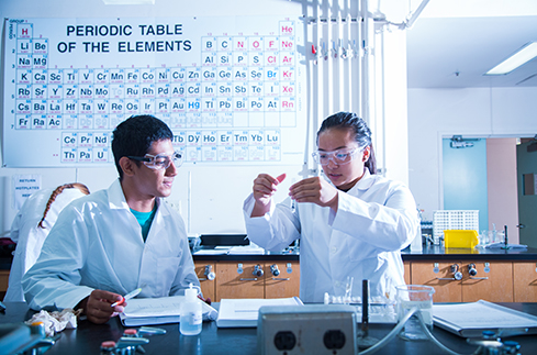 Image of two students in lab coats and goggles performing a lab