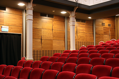 Image of a theater, indoors with red seats and wooden paneled walls 
